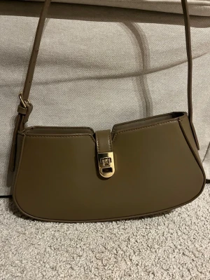 Olivgrön axelväska med gulddetalj - Snygg olivgrön axelväska i slätt konstläder med justerbar rem och guldfärgat spänne framtill. Väskan har en stilren, avlång form och minimalistisk design som passar till många olika outfits. 
