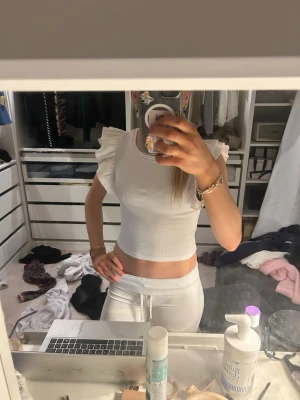 Vit croppad topp med volangärmar - Superfin vit croppad topp med volangdetaljer på axlarna. Toppen är ärmlös och har en tight passform som framhäver midjan. Perfekt för dig som gillar en söt och trendig stil. Materialet känns mjukt och stretchigt, troligtvis bomull eller en bomullsblandning.