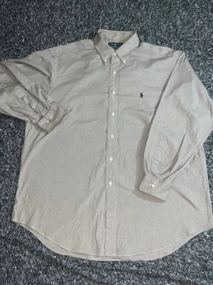 Beige skjorta från Ralph Lauren - Klassisk beige skjorta från Ralph Lauren med button-down krage och broderad logga på bröstet. Skjortan har långa ärmar, vita knappar och är tillverkad i bomull. Perfekt för en clean och stilren look. Skjortan är i bra skick och tveka inte på att höra av er vid intresse!