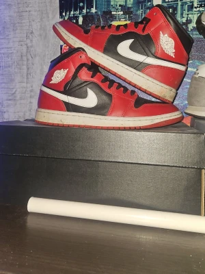 Nike Air Jordan 1 röd svart vit - Säljer ett par klassiska Nike Air Jordan 1 sneakers i rött, svart och vitt. Skorna har högt skaft, ikonisk swoosh-logga på sidan och Air Jordan-vingen på ankeln. Tillverkade i skinn med snörning framtill. Perfekta för dig som gillar streetwear och sneakerskultur. Skorna är ungefär helt nya använda cirka 10ggr små täcken på användning finns [smuts] inga som hälst skador på dem. Frågor?, Mer bilder tveka inte att slå en pling.