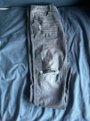 Grå slitna jeans Supply & Demand - Grå jeans från Supply & Demand med slitningar och hål på benen för en trendig, avslappnad look. Klassisk femficksmodell och knappgylf. Perfekta för dig som gillar streetwear och vill ha en edgy stil.