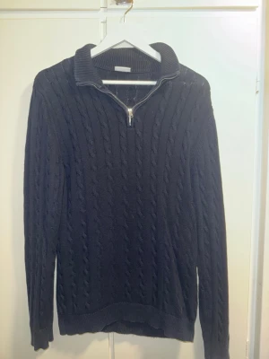 Mörkblå kabelstickad John Henric halv-zip - Snygg mörkblå kabelstickad tröja från John Henric med halv dragkedja och hög krage. Tröjan har långa ärmar och ribbade muddar vid ärmslut och nederkant. Perfekt för lager på lager och en klassisk och stilig look.