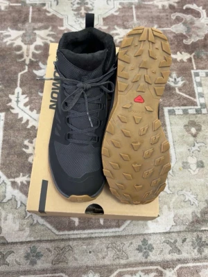 Svarta kängor från Salomon - Snygga svarta kängor från Salomon med robust gummisula i brunt och slitstark ovandel i textil och syntet. Skorna har snörning, vadderad insida och dragflik bak. Perfekta för utomhusaktiviteter och ger bra grepp på olika underlag.