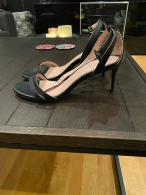 Svarta stilrena klackar med smal klack - Snygga svarta klackar med öppen tå och smal klack. Skorna har tunna remmar över tårna och runt ankeln med spänne. Insidan är ljusbeige och sulan är lätt vadderad för extra komfort. Perfekta för dig som gillar en klassisk och elegant look.