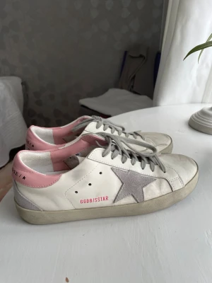 Golden Goose vita sneakers med rosa detalj - Golden Goose sneakers i vitt läder med grå mockadetaljer och en stjärna på sidan. Rosa hälparti med logotyp, grå skosnören och slitna detaljer för en trendig look. Perfekta för dig som gillar streetstyle och unika skor.