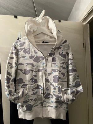 Grå camo zip hoodie från Bathing Ape - Säljer en grå camo zip hoodie från A Bathing Ape med hel dragkedja och fickor fram. Hoodien har ett unikt camomönster i grått, vitt och blått samt en stor huva med tryckt motiv. Perfekt för dig som gillar streetwear och vill sticka ut.