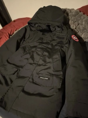 Svart parkas från Canada Goose - Säljer en svart parkas från Canada Goose med huva och stora fickor framtill. Jackan har klassisk patch på ärmen och stängs med både dragkedja och knappar. Perfekt för kalla dagar och riktigt snygg streetstil.