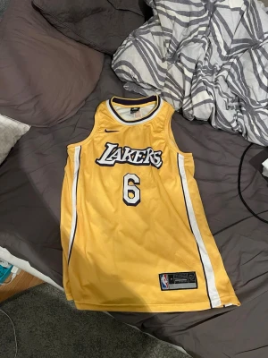 Gul Lakers basketlinne James #6 - Snyggt gult basketlinne från Nike med Lakers-logga och nummer 6 på framsidan samt 'James' och nummer 6 på ryggen. Klassisk NBA-design med vita och svarta detaljer längs sidorna. Perfekt för basketfans och streetwear.