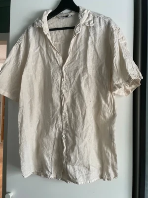 Beige linneskjorta från Justwest - Säljer en beige skjorta från Justwest i luftigt linne. Skjortan har korta ärmar, klassisk krage och knappar framtill. Perfekt för varma dagar och enkel att styla till olika outfits. Liten i storlek 