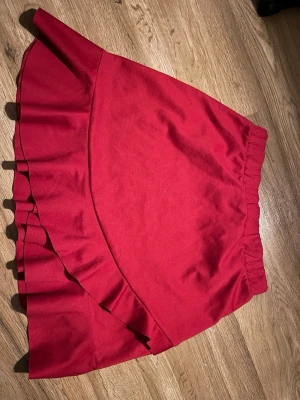 Röd volangkjol från Lindex - Supersnygg röd kjol från Lindex med bred resår i midjan och volangdetalj längs nederkanten. Kjolen har en enkel men trendig design och är gjord i mjukt bomullstyg som känns skönt mot huden.