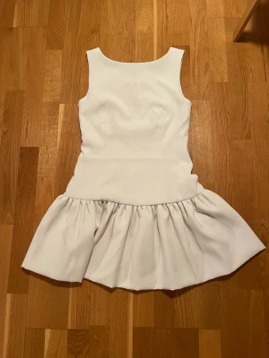 Vit kort klänning med volangkjol - Säljer en vit kort klänning med klassisk halsringning och breda axelband. Klänningen har en figurnära överdel och lite puff nedtill. Perfekt för sommarens alla tillfällen alt som studentklänning! Materialet känns mjukt och klänningen  är oanvänd. Säljs för att de blev fel storlek för mig.