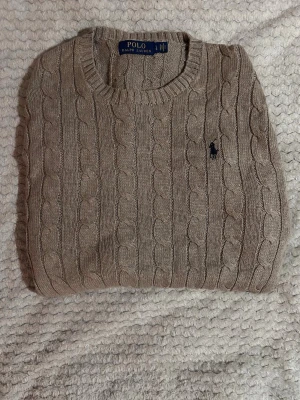 Beige/khaki kabelstickad tröja Polo Ralph Lauren - Kabelstickad beige/kaki tröja från Polo Ralph Lauren med rund halsringning och ribbade muddar. Tröjan har det klassiska broderade logotypen på bröstet och är tillverkad i ett mjukt material som känns lyxigt. Perfekt för dig som gillar tidlös och clean stil.