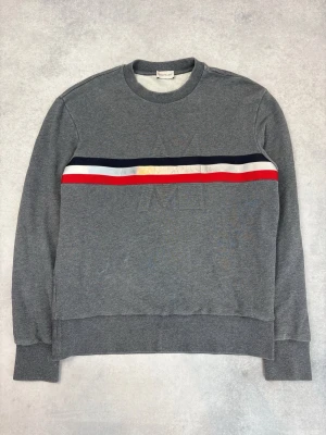 Grå sweatshirt från Moncler - Snygg grå sweatshirt från Moncler med rund hals och broderad logga på bröstet. Tröjan har en randig detalj i marinblått, vitt och rött över bröstet. Tillverkad i mjukt bomullsmaterial och har ribbade muddar vid ärmslut och nederkant. Perfekt för en avslappnad och stilren look.