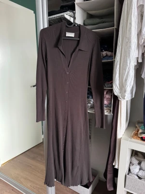 Brun ribbad klänning från Neo Noir - Mörkbrun ribbad klänning från Neo Noir med lång ärm, krage och knappar hela vägen framtill. Klänningen har en figurnära passform och är tillverkad i ett stretchigt, mjukt material som ger en snygg siluett.