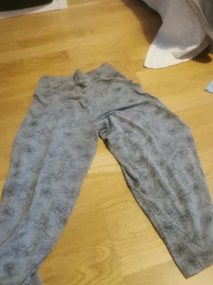 Grå mjukisbyxor med rosor - Grå mjukisbyxor med svart rosmönster över hela tyget. Byxorna har avslappnad passform och resår i midjan. Perfekta för chill dagar hemma. Materialet känns mjukt och bekvämt mot huden.