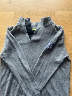 Grå stickad tröja från Salming - Snygg grå stickad tröja från Salming med hög krage och knappdetaljer vid halsen. Tröjan har ett broderat märke på ärmen med texten 'Salming Golf' och 'Authentic'. Perfekt för dig som gillar sportig stil och vill ha något bekvämt och stilrent. Original knapparna har ja tappat bort så jag har suttit dit egna