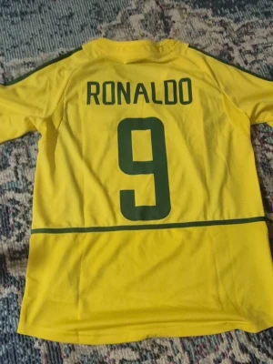 Ronaldo Nazario(R9) Brasilien hemmatröja - Säljer en klassisk gul Brasilien fotbollströja från Nike med gröna detaljer. Tröjan har nummer 9 och namnet Ronaldo på ryggen samt Brasilien-emblemet på bröstet. Perfekt för dig som älskar fotboll och vill bära legendarens tröja.