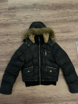 Svart pufferjacka med fuskpäls - Säljer en svart pufferjacka med avtagbar huva och fuskpäls. Jackan har flera dragkedjefickor, ribbade muddar och varm beige teddyfoder på insidan av huvan. Perfekt för kalla vinterdagar och riktigt snygg streetstil.