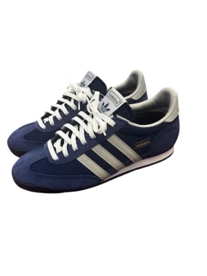 Blå Adidas sneakers med vita ränder - Snygga blå sneakers från Adidas med klassiska vita ränder på sidorna och vita skosnören. Ovandelen är i mocka och textil, och skorna har en låg profil med retrovibbar. Perfekta för dig som gillar stilrena och sportiga sneakers.