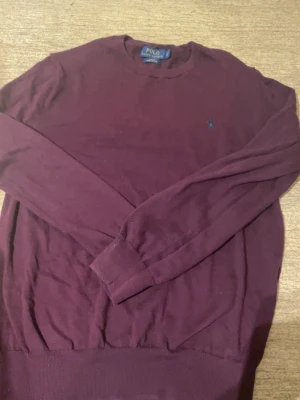 Vinröd stickad tröja Polo Ralph Lauren - Mysig vinröd stickad tröja från Polo Ralph Lauren med rund halsringning och diskret logga broderad på bröstet. Tröjan har långa ärmar och ribbade muddar vid ärmslut och nederkant. Perfekt för lager på lager och enkel att matcha.