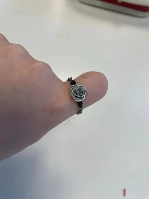 Silvrig ring med rosdetalj - Unik ring i silvrig metalltråd formad som en ros. Ringen har en flätad design runt hela bandet och rosdetaljen sitter i mitten. Perfekt för dig som gillar handgjorda och kreativa accessoarer med en cool twist.