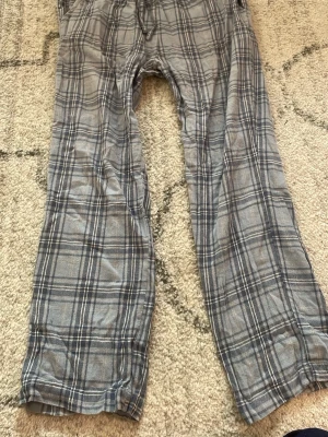 Grå rutiga pyjamasbyxor - Ett par grå pyjamasbyxor med klassiskt rutigt mönster i svart och vitt. Byxorna har resår i midjan och dragsko för justerbar passform. Perfekta för att chilla hemma eller sova i. Mjukt och bekvämt material.