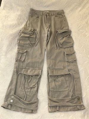 Grå cargobyxor med fickor - Säljer ett par beige cargobyxor med bred passform och flera stora fickor med lock och snörning. Byxorna har knappdetaljer vid benslut. Mycket bra skick, skriv vid frågor 🥰