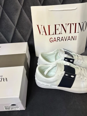 Valentino Garavani vita sneakers - Snygga vita sneakers från Valentino Garavani med bred svart rem över sidan och nitar på sulan. Skorna har vita skosnören, guldfärgad logga på plösen och är tillverkade i slätt läder. Perfekta för dig som gillar exklusiv och stilren design.