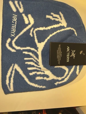 Blå mössa från Arc'teryx med logga - Snygg blå mössa från Arc'teryx med vit logotyp och grafiskt mönster på ena sidan. Tillverkad i mjukt stickat material som sitter skönt på huvudet. Perfekt för kalla dagar när du vill ha både stil och värme.