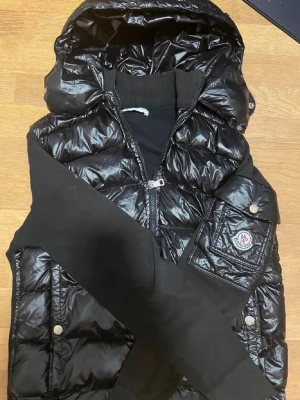Svart dunjacka från Moncler med huva - Snygg svart dunjacka från Moncler med glansig finish, huva och svarta stickade ärmar. Jackan har två framfickor med tryckknappar och Moncler-logga på ena ärmen. Perfekt för kalla vinterdagar och ger en sportig, trendig look. Vi kan snacka om priset 