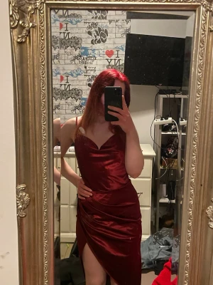 Vinröd långklänning med slits - Snygg vinröd långklänning i satin med smala axelband och v-ringning. Klänningen har en elegant slits framtill och figurnära passform som framhäver siluetten. Perfekt för dig som vill sticka ut med en lyxig look.