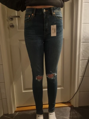 Blå high waist skinny jeans med hål - Säljer ett par blå high waist skinny jeans med slitna hål på båda knäna. Jeansen har klassiska fem fickor, gylf med dragkedja och knapp samt en tajt passform. Perfekta för dig som gillar en trendig och avslappnad stil. Aldrig använda.
