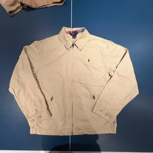 Beige jacka från Polo Ralph Lauren - Klassisk beige jacka från Polo Ralph Lauren med dragkedja framtill och diskret broderad logga på bröstet. Jackan har krage, långa ärmar med knappdetaljer vid ärmslut och två sidofickor. Perfekt för dig som gillar stilren och tidlös design.