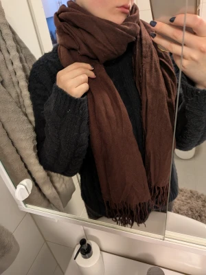 Mörkbrun halsduk med fransar - En stor och mysig mörkbrun halsduk med fransar i ändarna. Halsduken är rektangulär och tillverkad i ett mjukt tyg som känns skönt mot huden. Perfekt att vira flera varv runt halsen för extra värme.