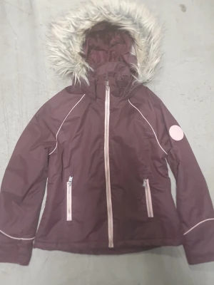 Vinterjacka - Snygg vinröd/lila vinterjacka med huva och fuskpälsdetalj. Jackan har vita dragkedjor och piping längs ärmarna samt två framfickor. Perfekt för kyliga dagar och har en modern, sportig look.