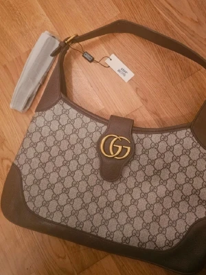 GG Supreme axelremsväska brun - Snygg axelremsväska i GG Supreme-mönstrad canvas med bruna läderdetaljer och guldfärgad GG-logga framtill. Väskan har en klassisk rektangulär form och bred axelrem. Perfekt för dig som gillar lyxiga accessoarer med ikonisk design.