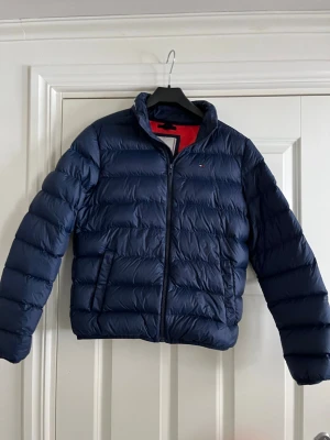 Mörkblå dunjacka från Tommy Hilfiger - Snygg mörkblå dunjacka från Tommy Hilfiger med quiltad design och dragkedja framtill. Jackan har ståkrage med logotyp och två fickor med dragkedja. Perfekt för kalla vinterdagar och har en sportig, stilren look.