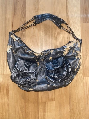 Silvrig axelremsväska med kedja - Cool handväska med två framfickor och detaljer i form av dubbla kedjor som axelrem. Väskan har en rundad form och stängs med dragkedja. 