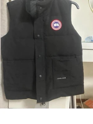 Svart dunväst från Canada Goose - Snygg svart dunväst från Canada Goose med klassisk logotyp på bröstet och stor ficka framtill. Västen har hög krage, tryckknappar och dragkedja. Perfekt för lager-på-lager och håller dig varm under kyliga dagar.