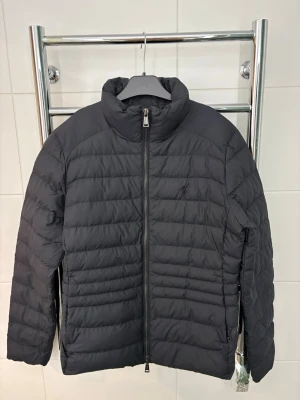 Svart pufferjacka från Ralph Lauren - Snygg svart pufferjacka från Ralph Lauren med diskret broderad logga på bröstet. Jackan har hög krage, dragkedja framtill och quiltad design. Perfekt för kalla dagar och enkel att matcha med olika outfits.