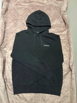 Svart hoodie från Peak Performance - Säljer en svart hoodie från Peak Performance med klassisk logga på bröstet. Hoodien har dragsko i huvan och en stor magficka. Tillverkad i mjukt bomullsmaterial och har ribbade muddar vid ärmslut och nederkant. Perfekt för en avslappnad och stilren look.