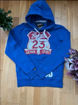 Blå Superdry hoodie med tryck - Snygg blå hoodie från Superdry med stort vitt och rött tryck på bröstet där det står 'Japan Spirit 23'. Klassisk huva med vita snören och ribbade muddar. Tillverkad i mjukt bomullsmaterial för en bekväm känsla. Perfekt för en avslappnad streetwear-look.