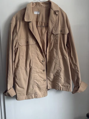 Beige overshirt från Tiger of Sweden - Stilren beige overshirt från Tiger of Sweden med klassisk krage, knäppning framtill och två bröstfickor med lock. Tillverkad i ett mjukt bomullstyg som ger en avslappnad men snygg look. Perfekt att bära öppen eller stängd över en t-shirt och suverän nu till våren.