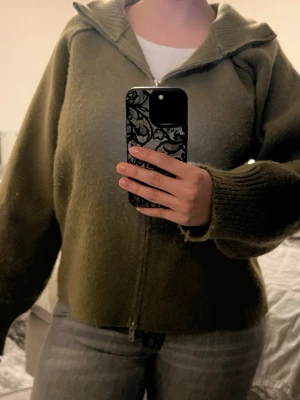 Olivgrön ziptröja med krage - Mysig olivgrön ziptröja med bred krage och långa ärmar. Tröjan har en rak passform och är tillverkad i ett mjukt, stickat material. Perfekt för lager på lager och enkel att matcha med jeans eller kjol.