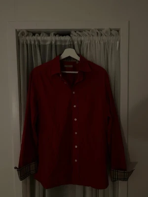 Röd skjorta från Burberry London - Snygg röd skjorta från Burberry med klassiskt rutigt mönster på insidan av kragen och manschetterna. Skjortan har långa ärmar, knappar framtill och en tidlös design som passar till många olika stilar. Tillverkad i bomull för en skön känsla.