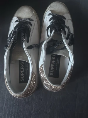Vita Golden Goose sneakers med glitter - Säljer ett par vita Golden Goose Super-Star sneakers med svart glittrig stjärna på sidan och guldglittrig häl. Skorna har svarta skosnören med text och är tillverkade i läder. Snygg och unik design som sticker ut.