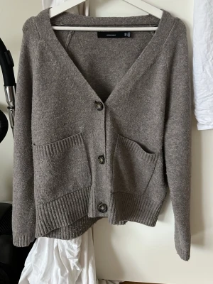Beige kofta från Vero Moda - Mysig beige kofta från Vero Moda med stora framfickor och knäppning framtill. Koftan har ribbstickade detaljer vid ärmslut och nederkant samt en avslappnad passform. Perfekt att slänga på sig när det är lite kyligt ute.