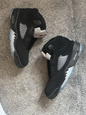Air Jordan 5 Retro Black Metallic - Säljer ett par Air Jordan 5 Retro Black Metallic sneakers. Skorna är svarta med silverdetaljer, meshpaneler på sidorna och en isblå sula. Tungan är silverfärgad med röd Jumpman-logga. Materialet är en mix av mocka och syntet. Klassisk high-top siluett.