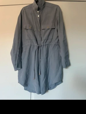Grå overshirt med knytband - Stilren grå overshirt med knappar framtill, två bröstfickor och knytband i midjan. Jackan har lång ärm och en rak, något längre passform. Perfekt för lager på lager och enkel att matcha med olika outfits.