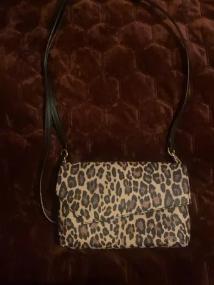 Leopardmönstrad axelväska - Snygg axelväska med leopardmönster i beige och brunt. Väskan har svart axelrem och guldfärgade detaljer. Perfekt för dig som vill sticka ut med ett djurmönstrat statement-plagg. Materialet ser ut att vara konstläder.